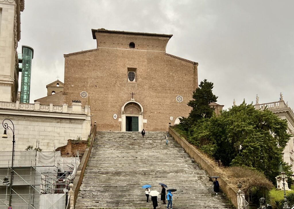 Escalinata empinada de piedra que conduce a la fachada de ladrillo de la Basílica de Santa Maria in Aracoeli en Roma, bajo un cielo nublado con personas subiendo con paraguas.