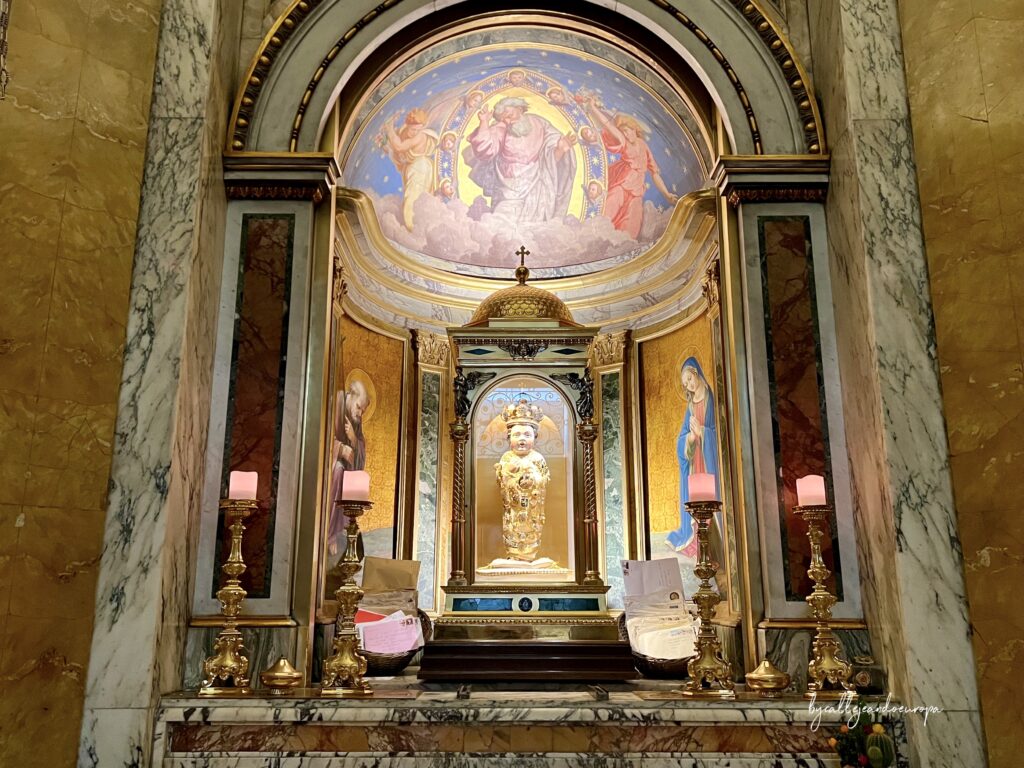 Altar ornamentado que alberga la figura del Santo Bambino, una pequeña estatua de madera del Niño Jesús cubierta de joyas y una corona de oro, situada en una hornacina con frescos de ángeles y figuras bíblicas al fondo.