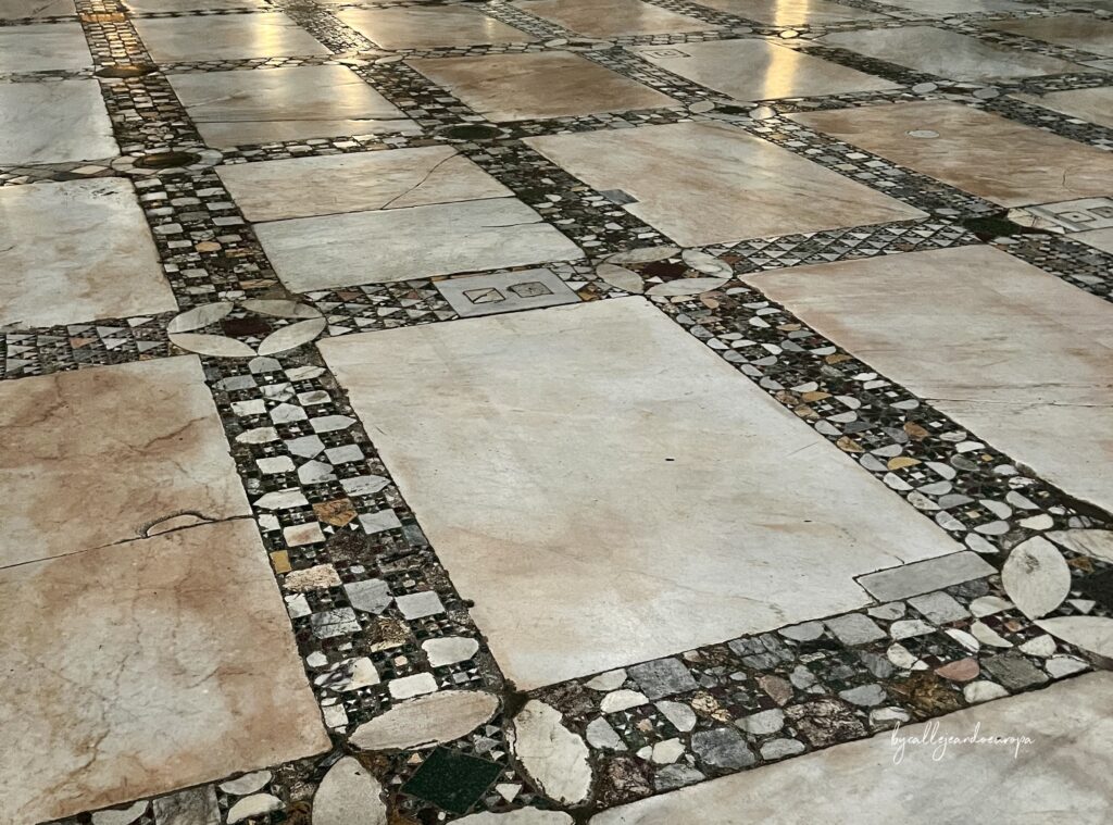 Primer plano de un suelo de estilo cosmatesco en la Basílica de Santa Maria in Aracoeli. Se aprecian grandes losas de mármol rodeadas por intrincados mosaicos geométricos hechos con pequeñas piezas de mármol de colores, pórfido y serpentina