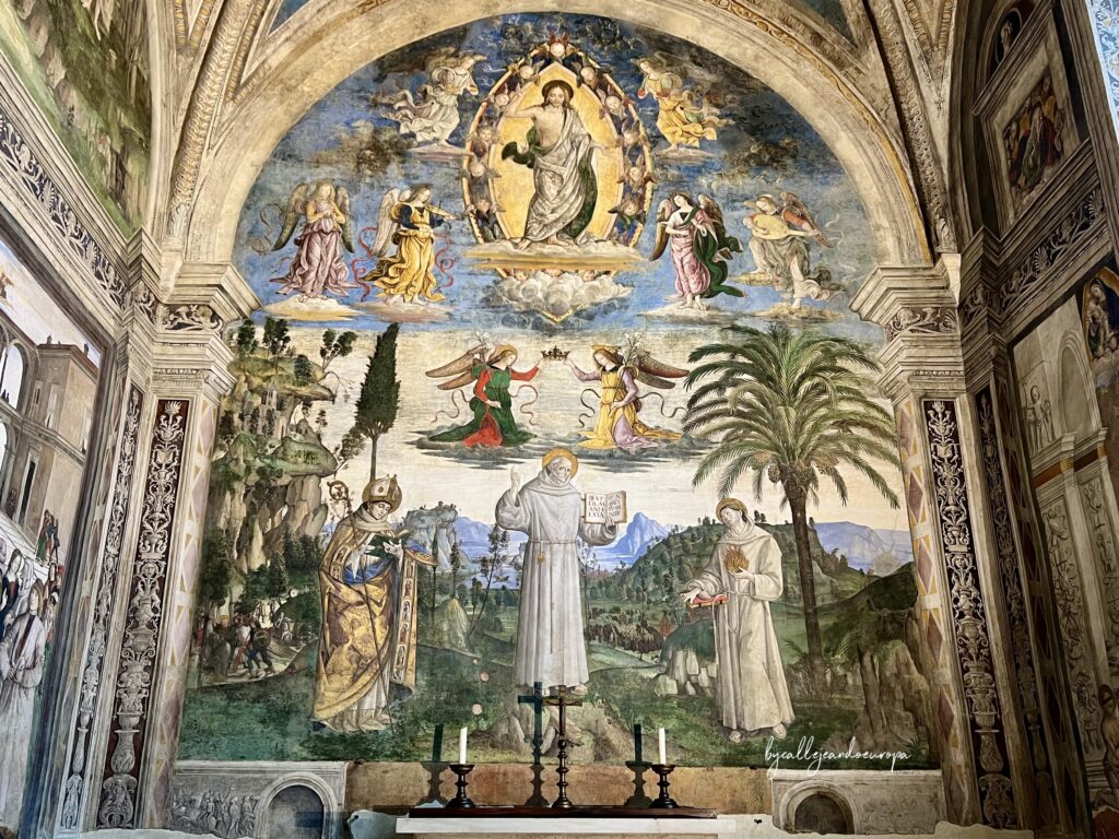 Fresco renacentista en una capilla abovedada que muestra a tres santos en un paisaje natural con una palmera; sobre ellos, ángeles sostienen una corona y, en la parte superior, Cristo en una mandorla rodeado de ángeles músicos.