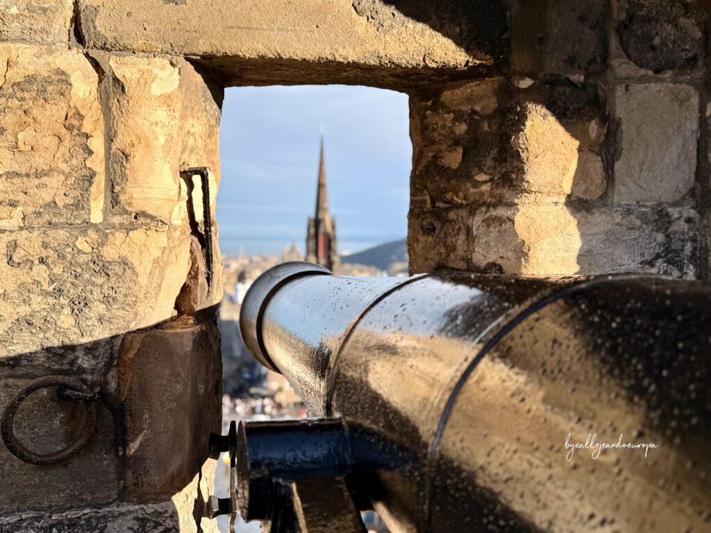 Primer plano de un cañón de hierro en el Castillo de Edimburgo apuntando hacia la ciudad, con la torre gótica de The Hub enmarcada al fondo