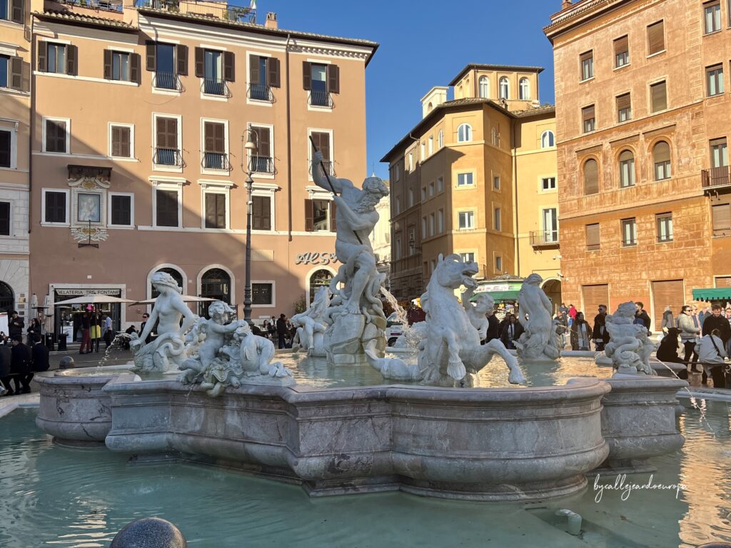 Vista de la Fuente de Neptuno en la Plaza Navona de Roma bajo un cielo despejado. La escultura central de Neptuno lucha con un pulpo, rodeado de estatuas de nereidas y caballos marinos en una gran pila de piedra. Al fondo, edificios clásicos de color ocre y personas paseando