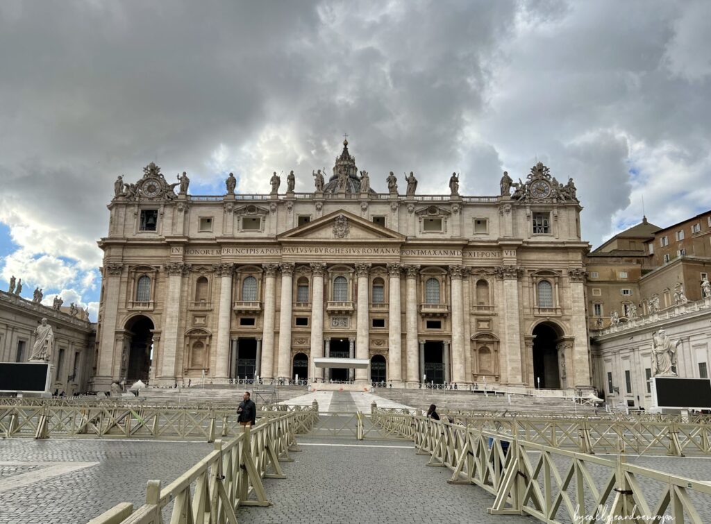 Fachada principal de la Basílica de San Pedro en el Vaticano bajo un cielo nublado, vista desde la Plaza de San Pedro con vallas de madera en primer plano para la audiencia papal.
