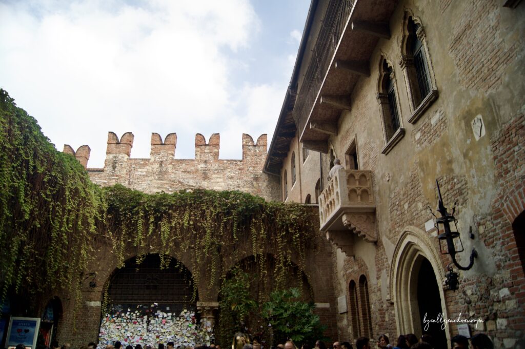 Vista del patio interior de la Casa de Julieta en Verona, mostrando el famoso balcón de piedra en una fachada de ladrillo, muros cubiertos de hiedra y una verja repleta de notas y candados de colores dejados por los visitantes.