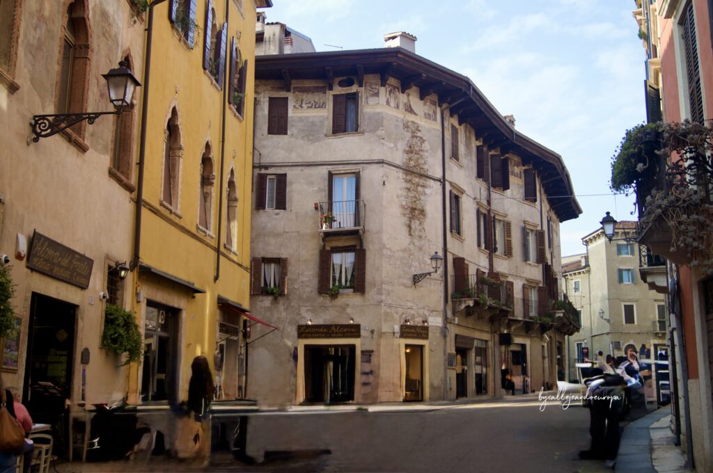 Vista de una calle histórica en Verona que conduce al Ponte Pietra, con edificios antiguos de fachadas ocres y crema, persianas de madera, balcones con flores y un restaurante llamado "Locanda Alcova" en la esquina.