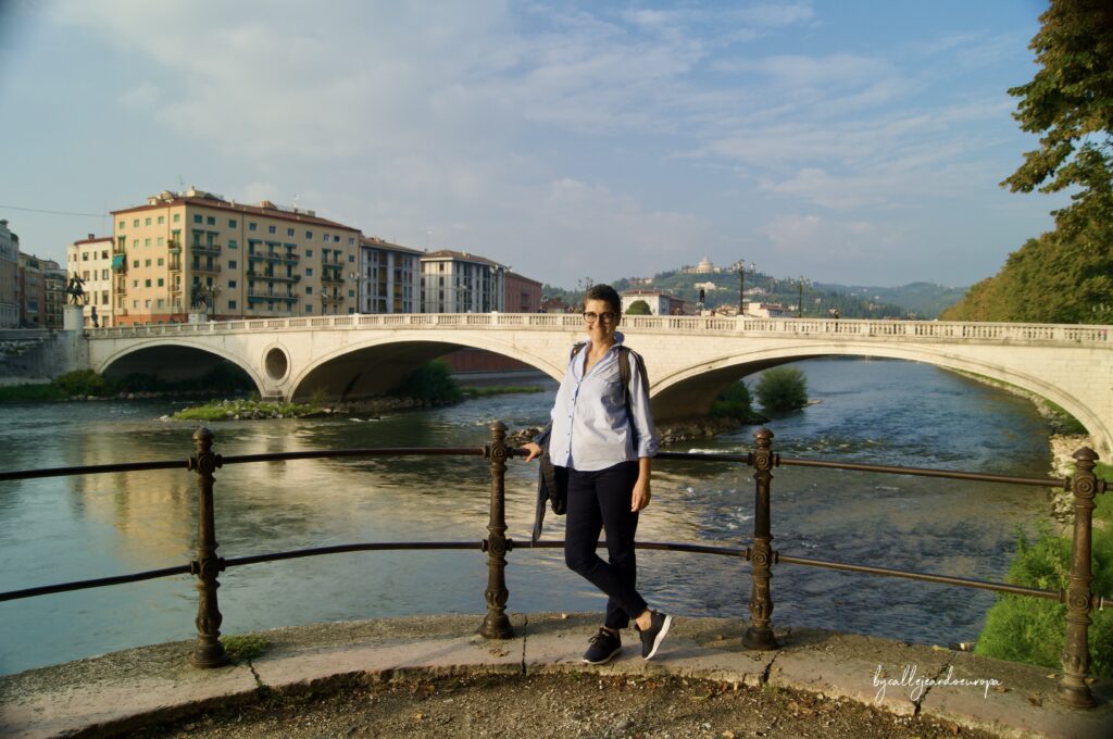 Callejeando Europa posa frente a una barandilla de hierro, con el Ponte della Vittoria y el río Adigio de Verona al fondo bajo un cielo despejado.