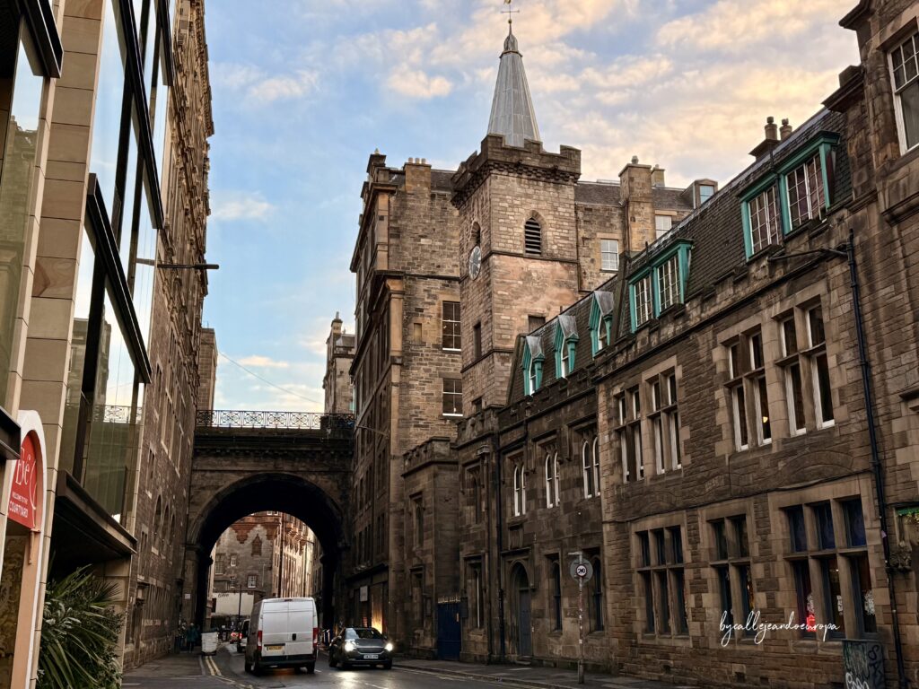 Vista de la calle Cowgate en Edimburgo, mostrando su arquitectura gótica de piedra oscura, un puente de arco antiguo cruzando la vía y una torre de reloj bajo un cielo atardecer.