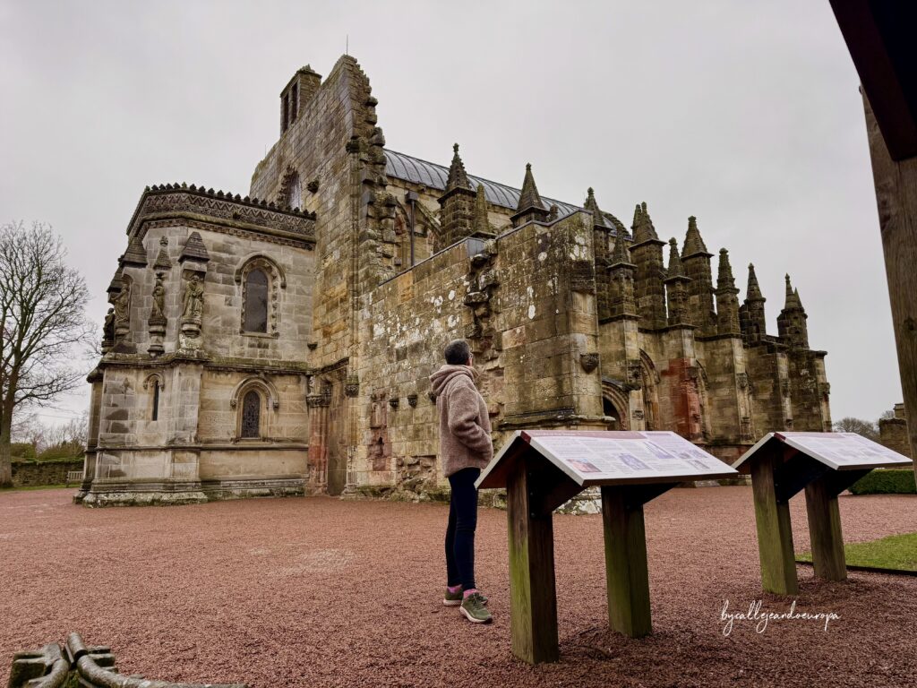 Vista exterior lateral de la Capilla de Rosslyn en un día nublado, con paneles informativos de madera en primer plano y Callejeando Europa observando la fachada.