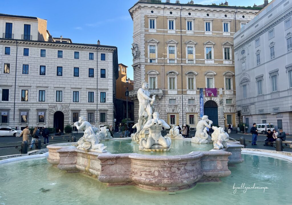 Vista frontal de la Fuente del Moro en la Piazza Navona, Roma. La escultura central representa a un etíope luchando con un delfín, rodeado por cuatro tritones que soplan caracolas. Al fondo se aprecian edificios históricos de arquitectura clásica bajo un cielo azul despejado