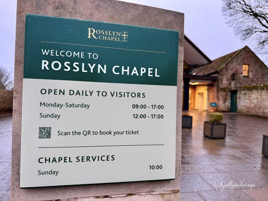 Cartel informativo en la entrada de Rosslyn Chapel con horarios de visita y código QR, con el edificio de piedra de fondo en un día lluvioso