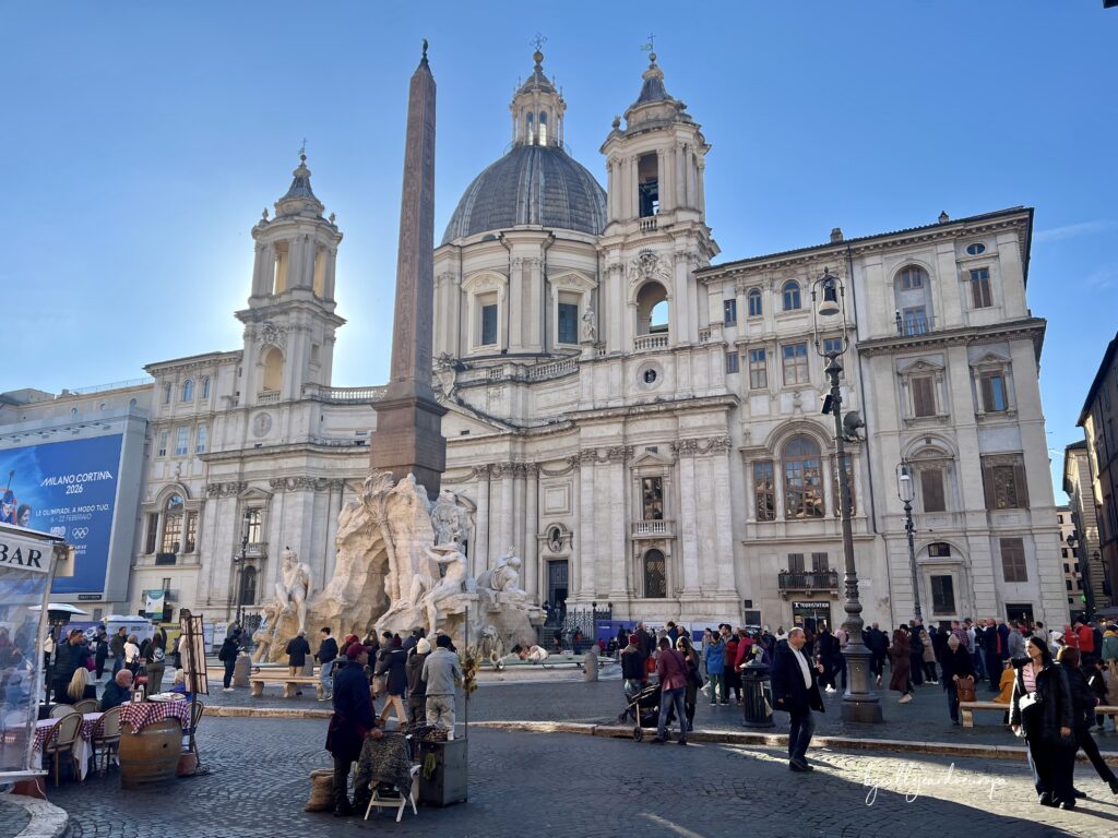 Fachada barroca de la Iglesia de Sant'Agnese in Agone en la Piazza Navona, destacando su gran cúpula central y sus dos torres campanario laterales. En primer plano se observa la Fuente de los Cuatro Ríos con su obelisco egipcio, rodeada de transeúntes en un día soleado en Roma.