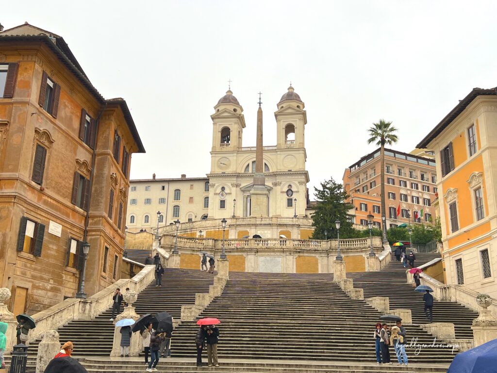 La escalinata de la Plaza de España en Roma subiendo hacia la iglesia de Trinità dei Monti, con turistas paseando por los peldaños de piedra.
