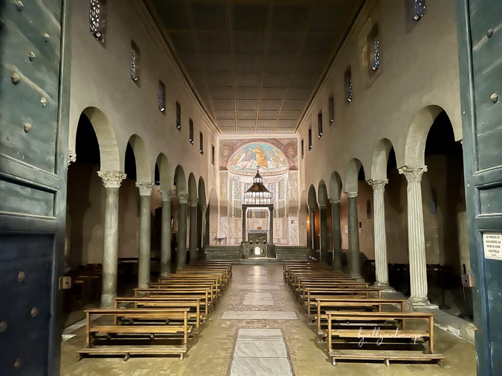 Vista del interior de la Iglesia de San Giorgio al Velabro en Roma, mostrando la nave central flanqueada por columnas antiguas y dos naves laterales, con bancos de madera y un fresco en el ábside al fondo.