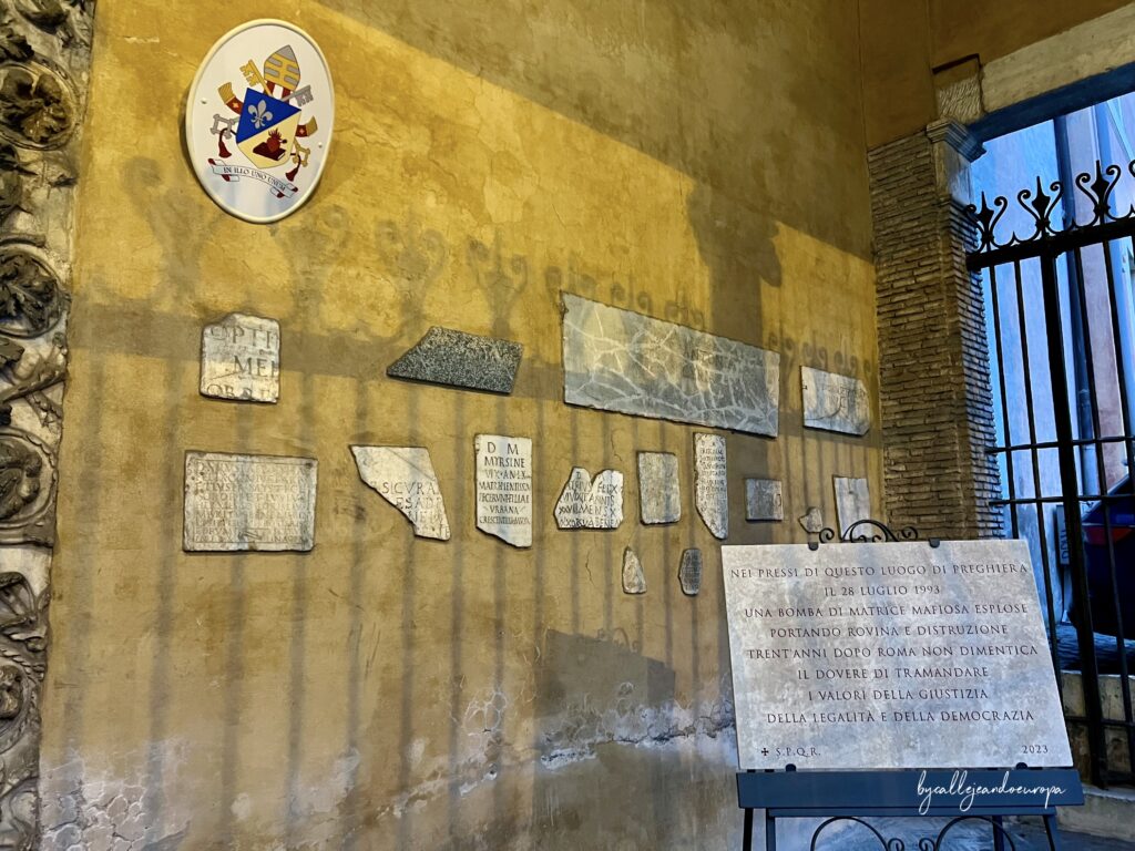 Primer plano de una pared lateral en San Giorgio al Velabro con fragmentos de lápidas antiguas de mármol incrustadas. A la derecha, una placa moderna conmemora el atentado de 1993, situada bajo un escudo heráldico papal en una pared de tono ocre.