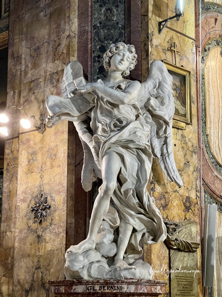 Escultura barroca de mármol blanco de un ángel con expresión de duelo, sosteniendo el pergamino del INRI (Título de la Cruz). La obra, de Gian Lorenzo Bernini, está situada sobre un pedestal de mármol veteado frente a una columna decorada