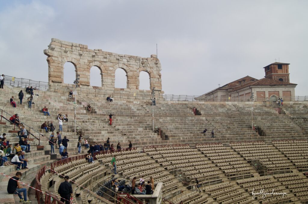 Vista del interior de la Arena de Verona con las gradas de piedra originales, visitantes sentados y la icónica "Ala" (fragmento del anillo exterior) destacando contra el cielo.