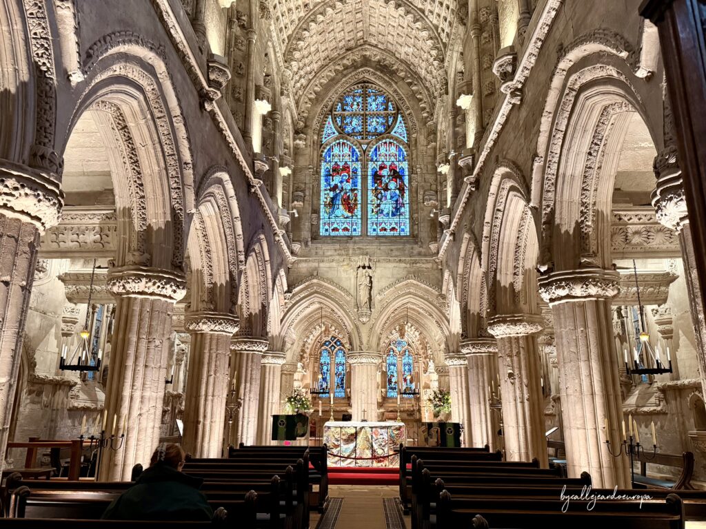 Vista del altar y las naves de Rosslyn Chapel, destacando los intrincados tallados en piedra de los arcos y las coloridas vidrieras iluminadas.