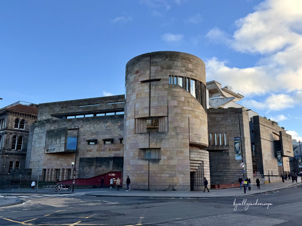 Vista exterior del Museo Nacional de Escocia en Edimburgo. Destaca su moderna torre circular de piedra arenisca con aberturas geométricas y ventanales verticales.