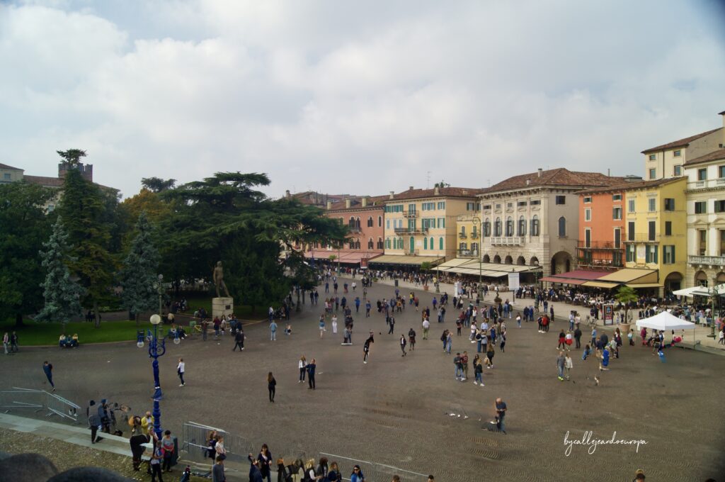 Vista panorámica de la Piazza Bra en Verona desde una posición elevada, mostrando la amplia plaza adoquinada con peatones, edificios de colores vibrantes con terrazas de restaurantes y árboles frondosos a la izquierda.