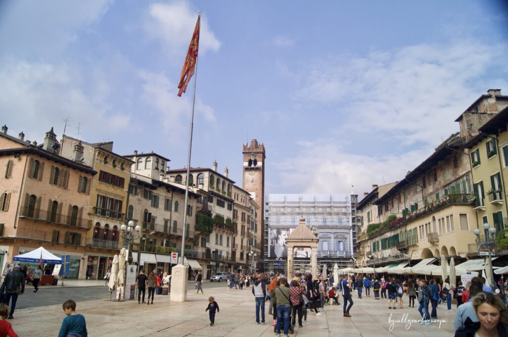 Vista amplia de la Piazza delle Erbe en Verona, destacando la Torre dei Lamberti al fondo, edificios históricos con fachadas decoradas y una gran bandera roja y dorada ondeando en un mástil frente a una multitud de personas.