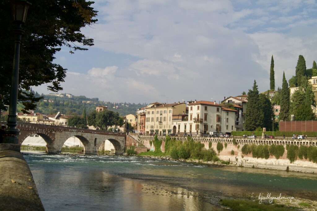 Vista del Puente de Piedra romano cruzando el río Adigio en Verona, con edificios históricos en la orilla, colinas verdes al fondo y el reflejo de las estructuras en el agua bajo un cielo parcialmente nublado.