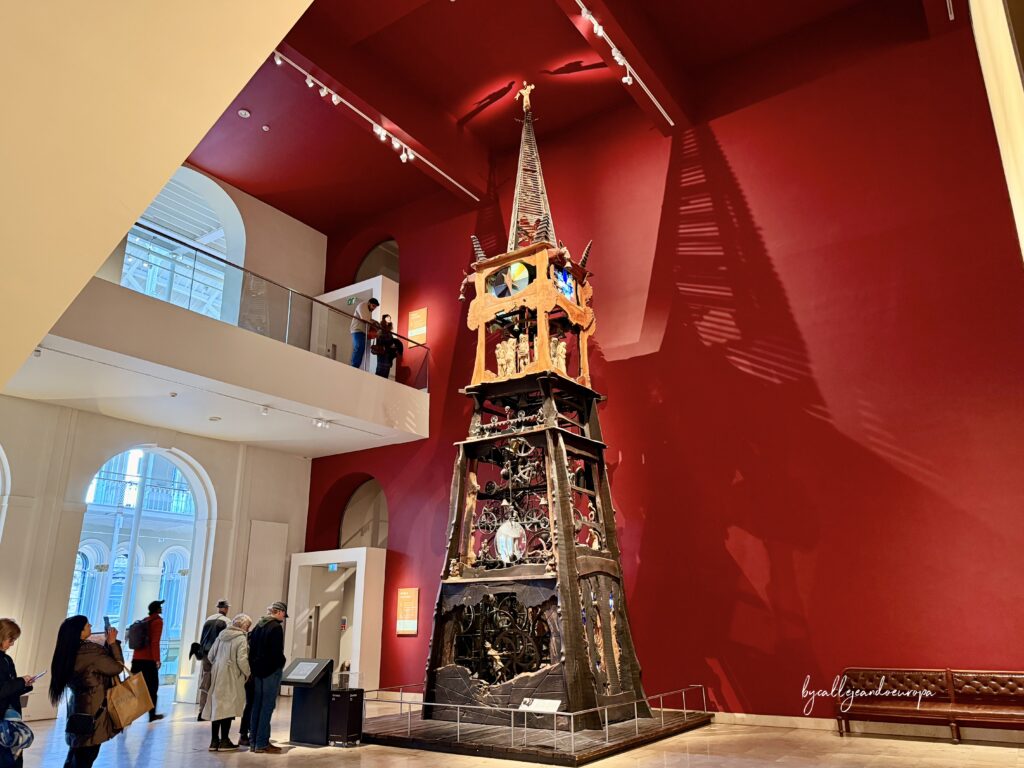 Una alta y compleja torre de reloj de madera y metal, conocida como el Reloj del Milenio, situada en una sala de museo con una gran pared de fondo color rojo intenso. La estructura muestra engranajes expuestos, figuras talladas y una aguja gótica en la cima.