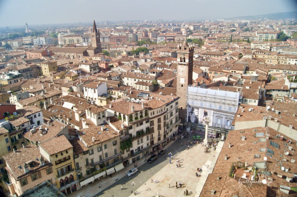Vista aérea en picado de la Piazza delle Erbe en Verona, mostrando los tejados de terracota, la plaza llena de gente, la Columna del León de San Marcos y la Torre del Gardello junto al Palacio Maffei.