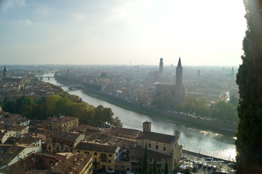 Vista panorámica de la ciudad de Verona desde un mirador, mostrando el río Adigio serpenteando entre edificios históricos, puentes y torres de iglesias bajo una luz suave de mañana.