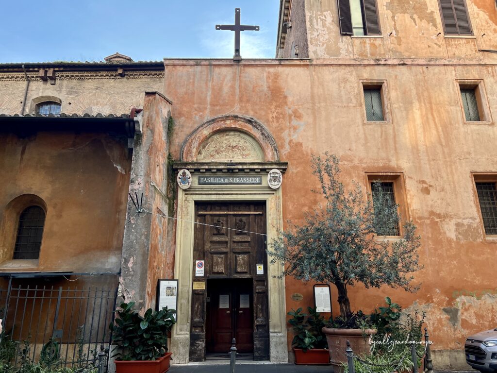Fachada exterior de la Basílica de Santa Práxedes en Roma, mostrando la puerta principal de madera bajo un arco de medio punto con una cruz en la parte superior