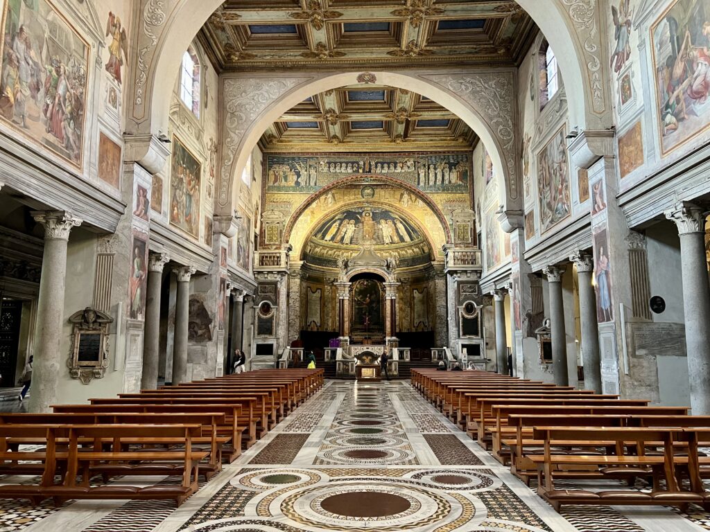 Vista de la nave central de la Basílica de Santa Práxedes en Roma, destacando el suelo de estilo cosmatesco, los bancos de madera y los mosaicos bizantinos del arco triunfal y el ápside