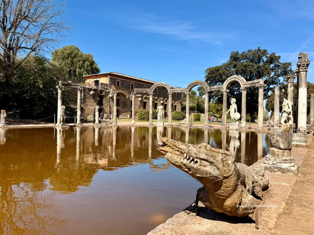 Escultura de cocodrilo en primer plano junto al estanque del Canopo con reflejos de columnas y arcos romanos en Villa Adriana.