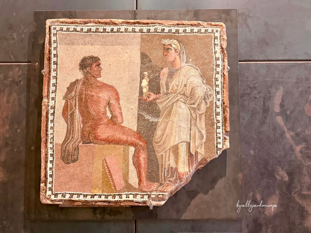 Mosaico antiguo conservado en los Museos Capitolinos que representa a un hombre sentado de espaldas y a una figura femenina de pie portando una estatuilla.