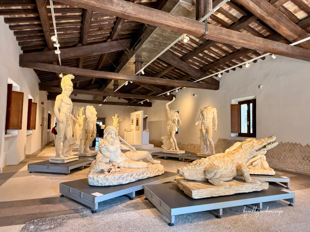Sala del museo Antiquarium de Villa Adriana con estatuas clásicas de mármol y un cocodrilo esculpido bajo un techo de vigas de madera.