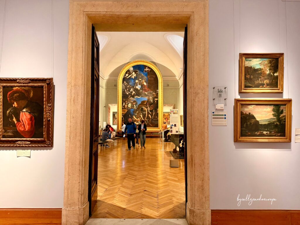 Vista a través de una puerta de la Sala IV de la Galería Borghese en Roma, mostrando varias pinturas clásicas colgadas en las paredes y visitantes en el interior.
