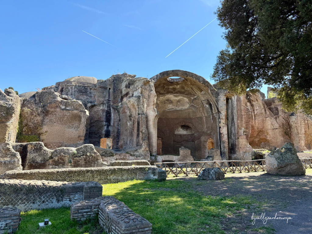 Vista exterior de las monumentales ruinas de las Termas en Villa Adriana, mostrando la gran bóveda semicircular de ladrillo bajo un cielo azul con rastros de aviones.